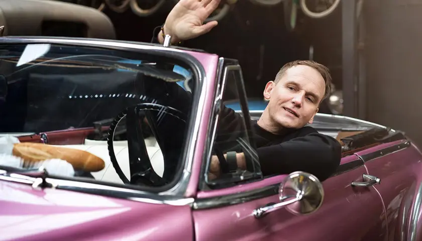 Pascal Brodnicki macha ręką z różowego kabrioletu. W samochodzie bagietka i kierownica.