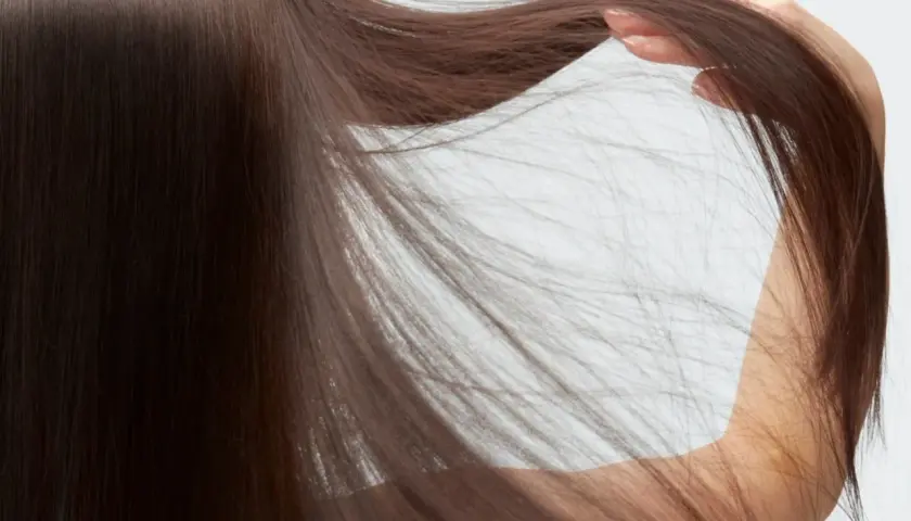 Mano acariciando cabello castaño, fino y escaso, que cae en cascada sobre un fondo blanco.