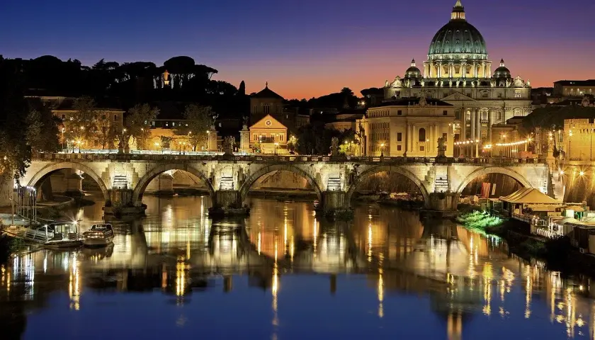 Ponte Sant'Angelo illuminato di notte, con la cupola di San Pietro sullo sfondo. Un'idea per cosa visitare a Roma in due giorni.