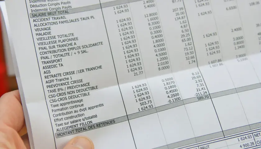 Une main tient une fiche de paie perdue, détaillant les salaires et les retenues.