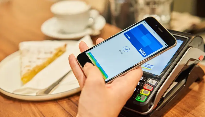 Bezahlvorgang mit Apple Pay: Ein Smartphone hält über einem Kartenlesegerät, daneben ein Stück Kuchen.