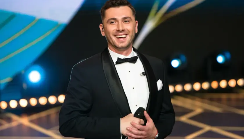 Kamil Bednarek w czarnym smokingu z muszką, trzyma mikrofon i uśmiecha się.