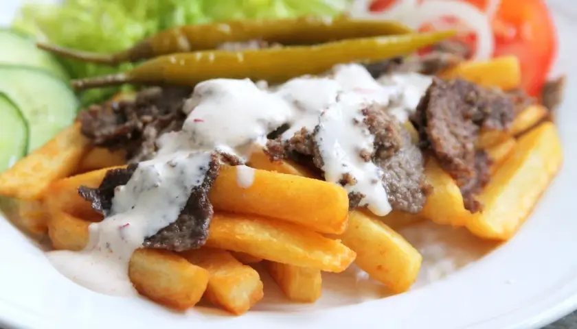 En tallrik med pommes frites, kebabkött och vit sås. Tillbehör som sallad, gurka, tomat, lök och jalapeños. Ett gott kebab recept!