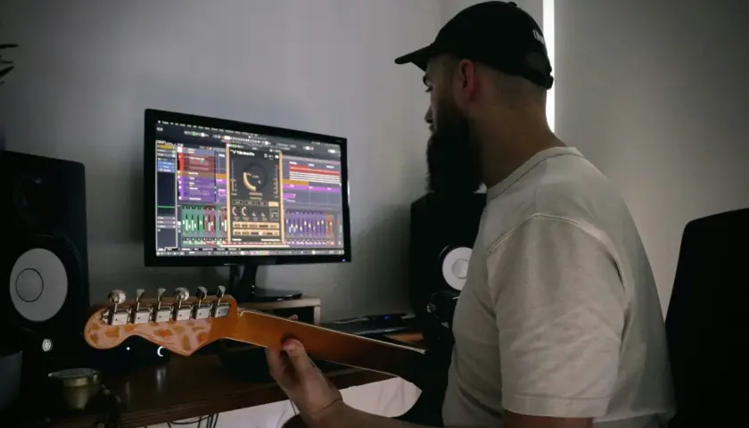 Mężczyzna z brodą i czapką z daszkiem uczy się, jak podłączyć gitarę do PC, pracując nad muzyką na komputerze.