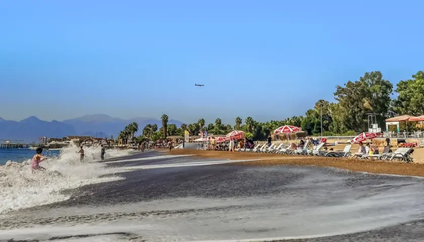 Ein sonniger Antalya Strand mit sanften Wellen, Liegestühlen und Palmen. Ein Flugzeug zieht seine Bahn am blauen Himmel.