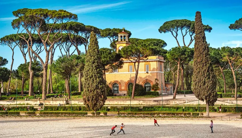 Un edificio giallo con una torre dell'orologio, circondato da pini marittimi e cipressi. Bambini giocano in un'area sabbiosa. Ecco cosa vedere ai Parioli.