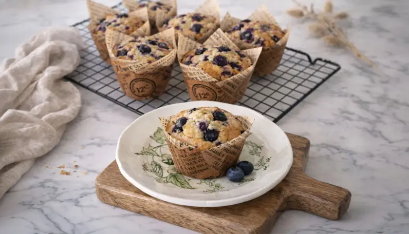 Un délicieux muffin myrtille, tout juste sorti du four, posé sur une assiette décorée. D'autres muffins myrtilles refroidissent sur une grille.