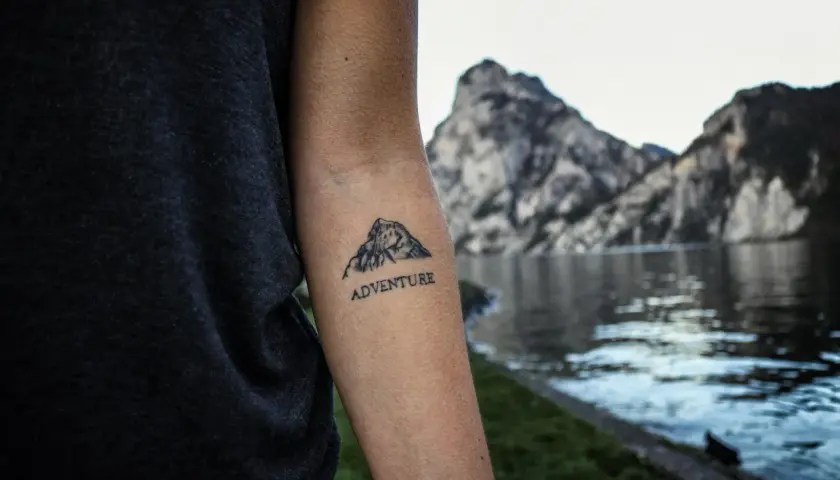 Tatuaż góry z napisem "ADVENTURE" na przedramieniu. W tle jezioro i skaliste szczyty.