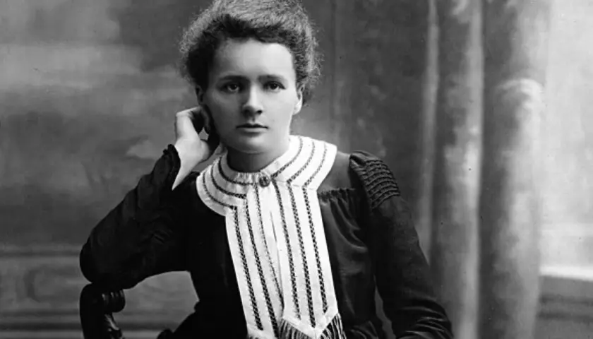 Czarno-białe zdjęcie Marii Skłodowskiej-Curie w eleganckiej sukni z ozdobnym kołnierzem.