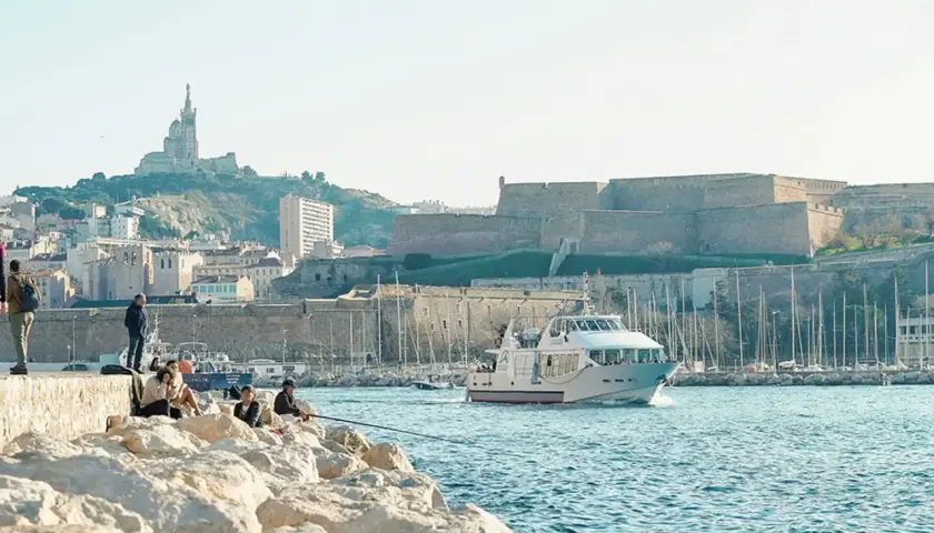 Widok na port w Marsylii z łodzią, fortecą i bazyliką na wzgórzu.