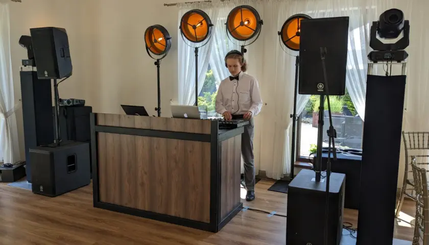 Młody DJ w muszce i białej koszuli obsługuje sprzęt muzyczny przy drewnianym stanowisku, otoczony głośnikami i oświetleniem.