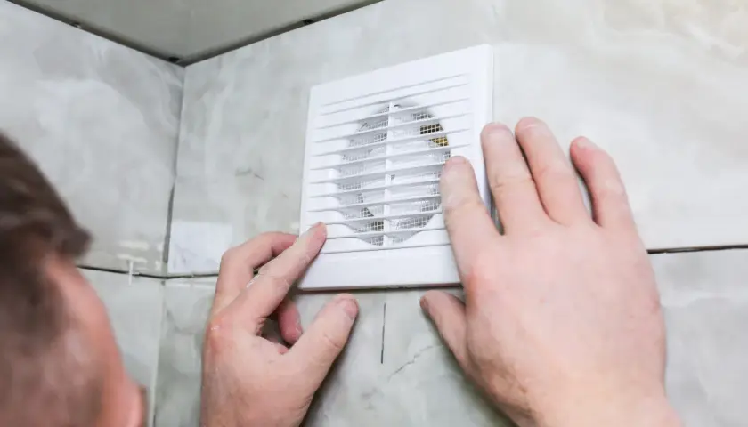 Installation d'un ventilateur pour l'aération de la salle de bain. Des mains ajustent l'appareil blanc sur un mur carrelé.