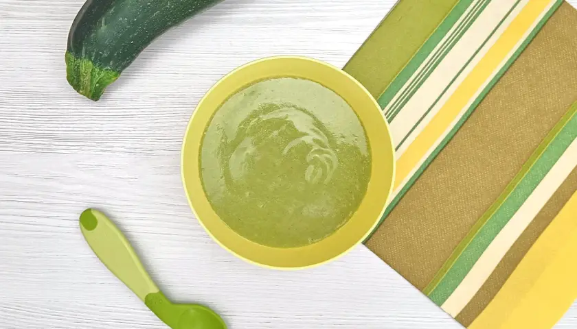 Un plato con puré de calabacín bebé, una cuchara verde y un calabacín entero sobre una mesa de madera.