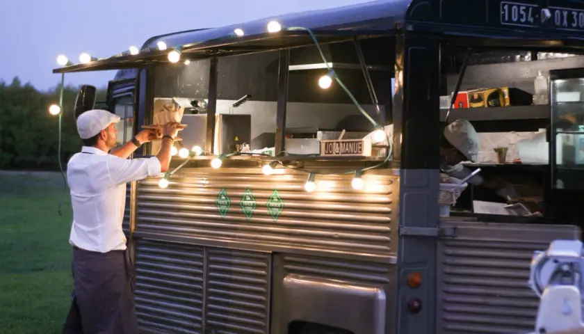 Un food truck illuminé de guirlandes lumineuses, parfait pour une idée food truck mariage. Un serveur sert un plat.