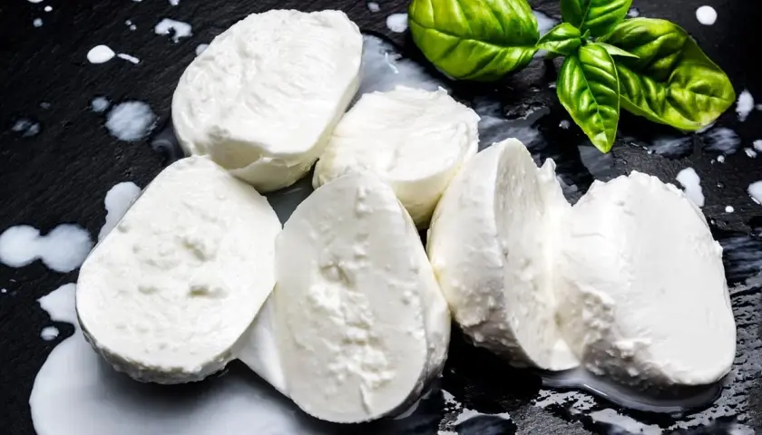 Świeża mozzarella na czarnym tle, z kilkoma listkami bazylii. Czy mozzarella jest kaloryczna? To zależy od jej rodzaju, ale zazwyczaj jest umiarkowanie kaloryczna.