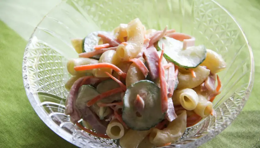 A close-up of a crystal bowl filled with creamy Japanese macaroni salad, featuring elbow macaroni, shredded carrots, sliced cucumbers, and ham.