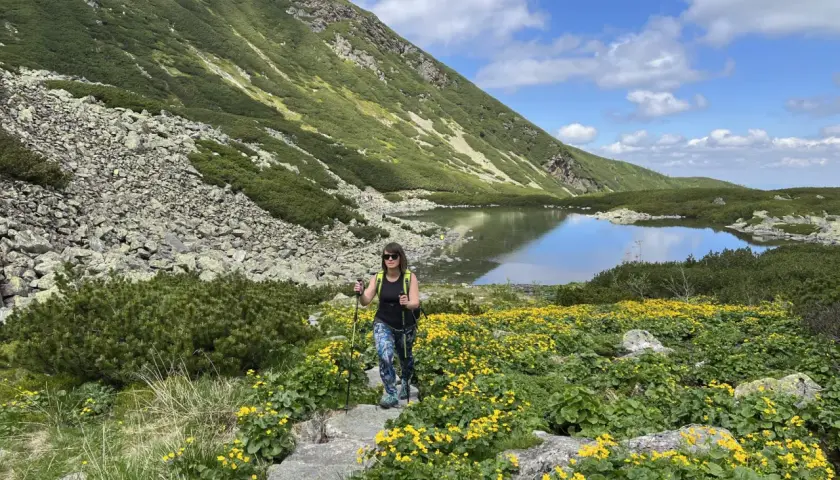 Kobieta z kijkami trekkingowymi idzie przez kwitnącą łąkę w górach. To najłatwiejszy szlak na Krzyżne, z jeziorem w tle.