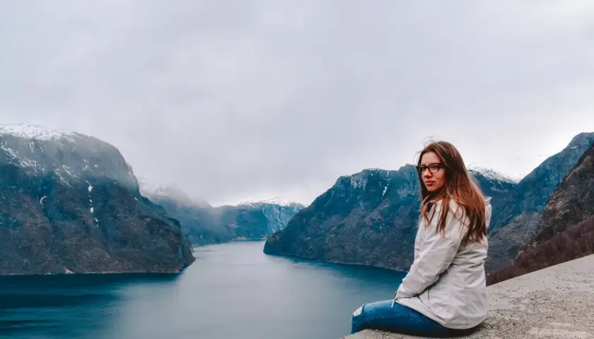 Kobieta siedzi na skarpie z widokiem na norweski fiord i ośnieżone góry.