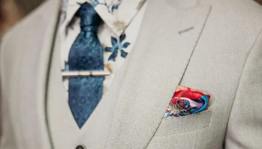 Elegancki mężczyzna w jasnej marynarce, z niebieskim wzorzystym krawatem i spinką do krawata jak nosić. W kieszeni kolorowa poszetka.