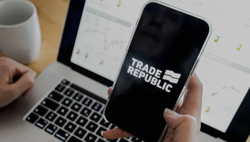 Person prüft den Trade Republic Quartalsbericht auf dem Smartphone, während der Laptop mit Finanzdiagrammen im Hintergrund läuft.