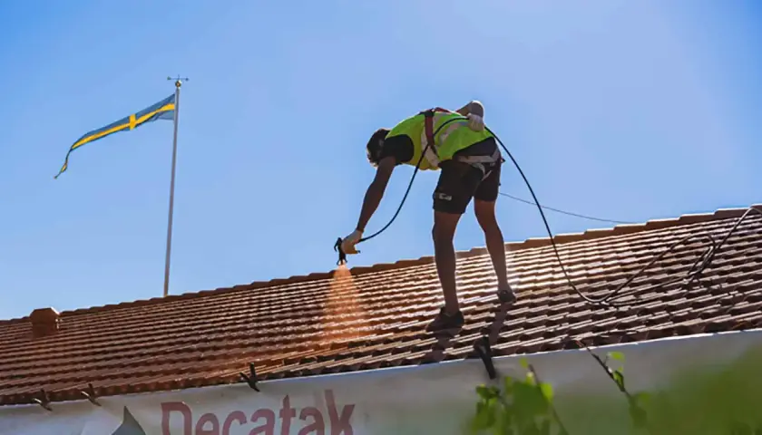 Arbetare sprutmålar tak med svensk flagga i bakgrunden.