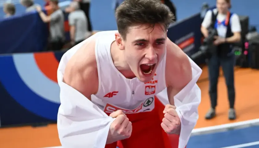 Młody lekkoatleta w biało-czerwonej koszulce okryty białą flagą, krzyczy z radości po zwycięstwie na halowych mistrzostwach europy lekkoatletyka.