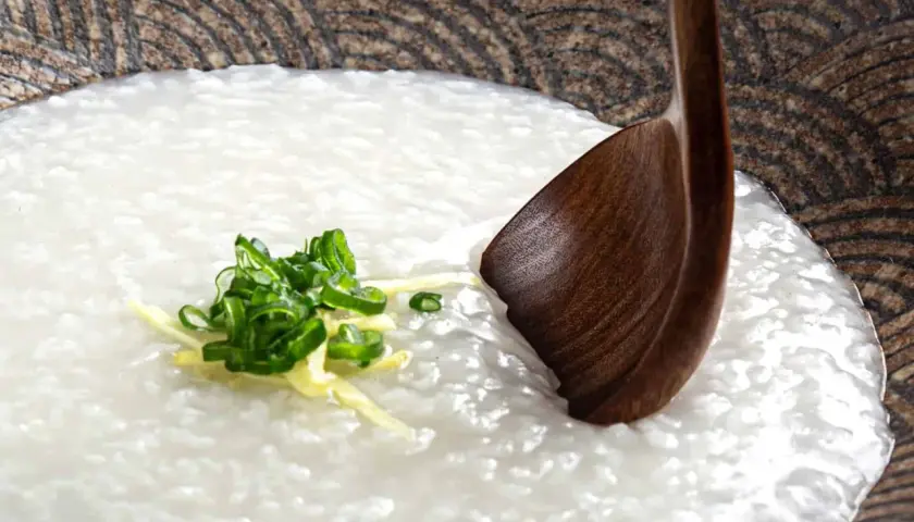 A comforting bowl of Japanese rice porridge (okayu) garnished with fresh scallions and ginger, ready to be enjoyed with a wooden spoon.