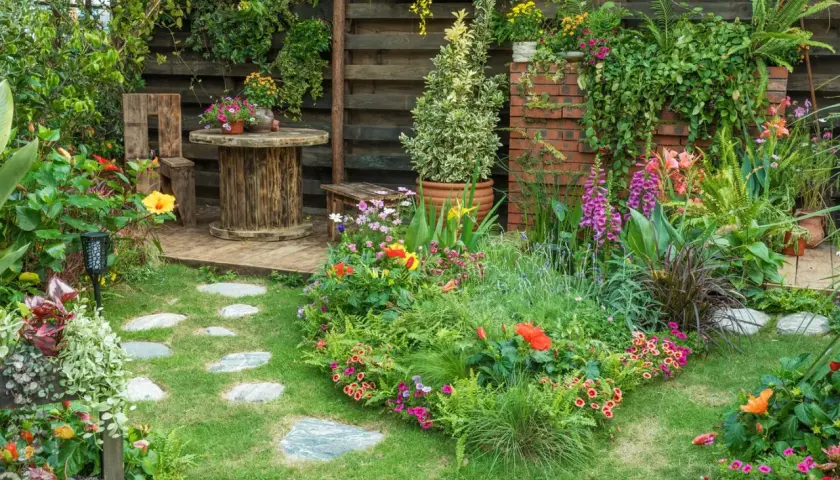 Ein üppiger, naturnaher Garten mit bunten Blumen, grünen Pflanzen und einem rustikalen Sitzbereich. Steinplatten führen durch das satte Grün.
