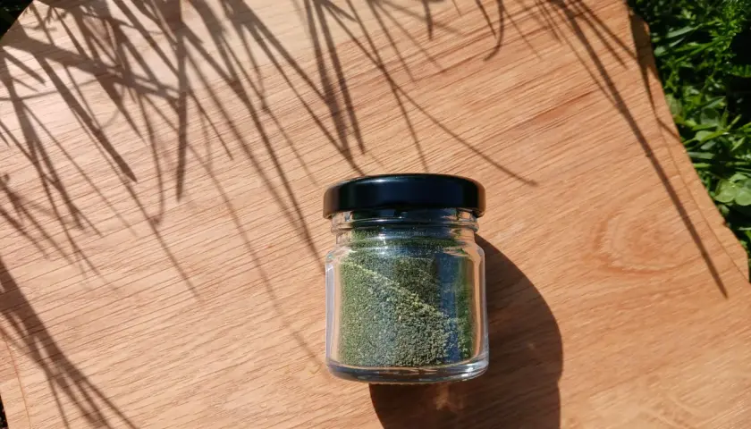Un piccolo barattolo di vetro contiene polvere di ortica verde brillante, posato su un tagliere di legno con ombre di foglie.
