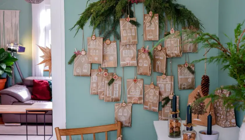 Selbstgemachter Adventskalender für Erwachsene: Kleine Papiertüten mit Hausmotiven und Zahlen hängen an Tannenzweigen.