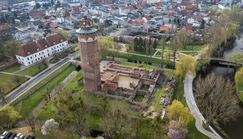 Ruiny zamku krzyżackiego w Brodnicy z charakterystyczną wieżą, otoczone zielenią i wodą, w tle rozciąga się miasto.