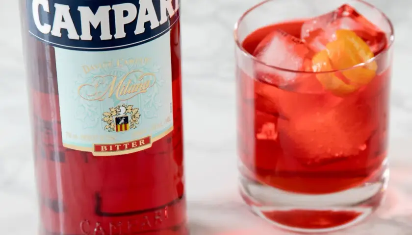 Butelka Campari i szklanka z czerwonym drinkiem, lodem i skórką pomarańczy. Campari to gorzki aperitif.