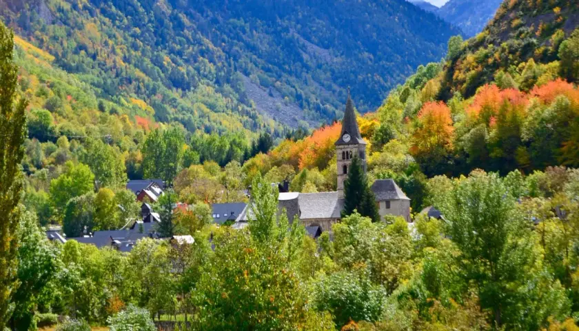 Pueblo en el **Valle de Arán** rodeado de árboles con hojas de otoño en tonos verdes, amarillos y naranjas. Destaca una iglesia con campanario.
