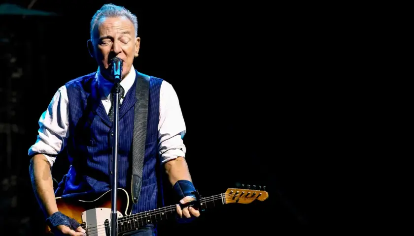 Bruce Springsteen z gitarą na scenie, w niebieskiej kamizelce i białej koszuli. Niezapomniane koncerty legendy rocka.