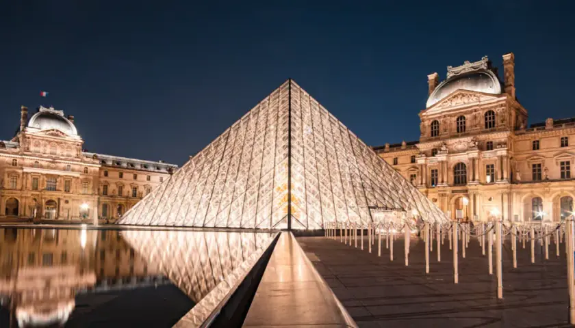 Piramide Luwru nocą z odbiciem w wodzie i oświetlonymi budynkami muzeum.