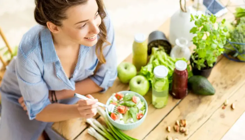 Junge Frau isst gesunden Salat. Viele gesunde Lebensmittel wie Obst, Gemüse und Nüsse sind auf dem Tisch, die reich an Cystein sind.