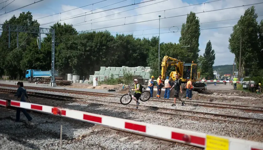 Pracownicy budowlani i dróżnik przejazdowy nadzorują prace przy torach. Koparka i ciężarówka są na miejscu.