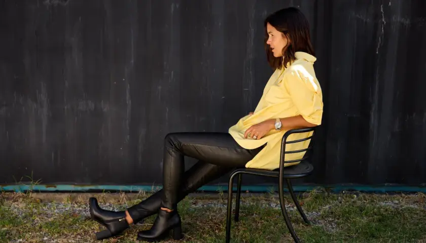 Une femme assise porte un pantalon avec bottines homme, une chemise jaune vif et une montre.