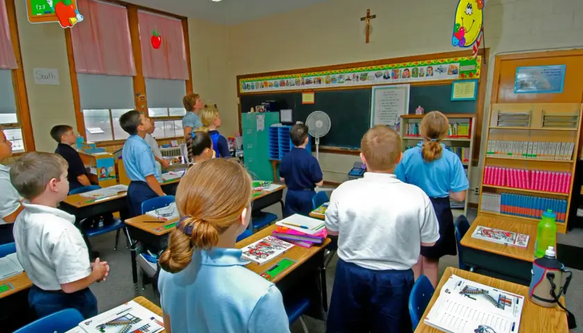 Dzieci słuchają katechety w klasie. Na ścianie wisi krzyż i rysunki.