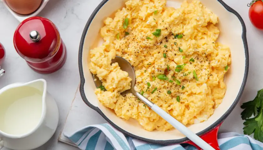 Oeufs brouillés crémeux, parsemés de ciboule fraîche et de poivre noir, servis dans une poêle rouge. Un délice pour le petit-déjeuner.