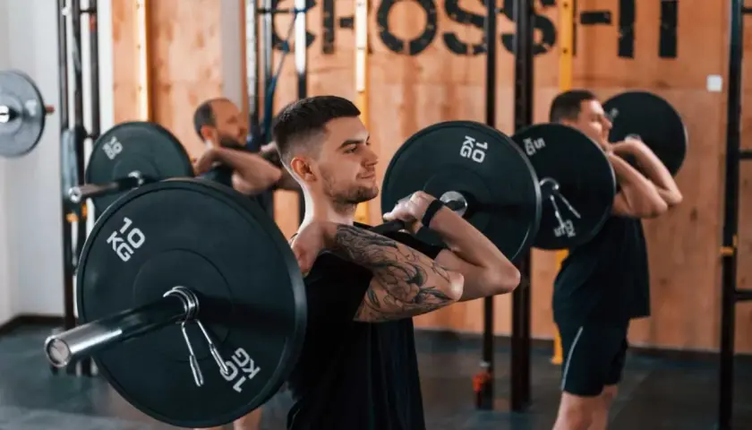 Trzech mężczyzn w siłowni CrossFit wykonuje ćwiczenia ze sztangą na klatce piersiowej.