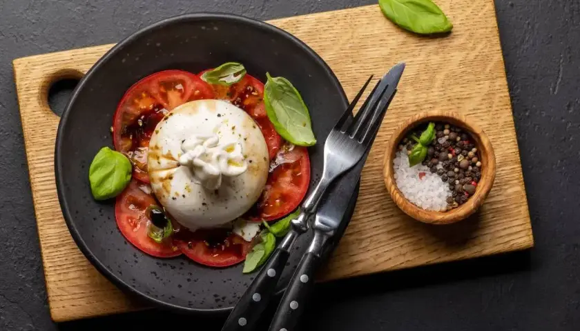Pyszna burrata z pomidorami, polana oliwą, ozdobiona bazylią. Obok miseczka z solą i pieprzem.