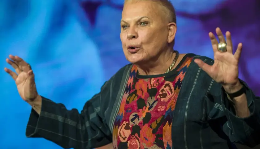 Elżbieta Dzikowska w kolorowej bluzce z haftem, gestykuluje rękami na tle niebieskiego ekranu.