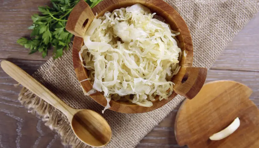 Holzschale mit Sauerkraut, daneben ein Holzlöffel und ein Stück Knoblauch. Manche finden, dass Sauerkraut schmeckt bitter.