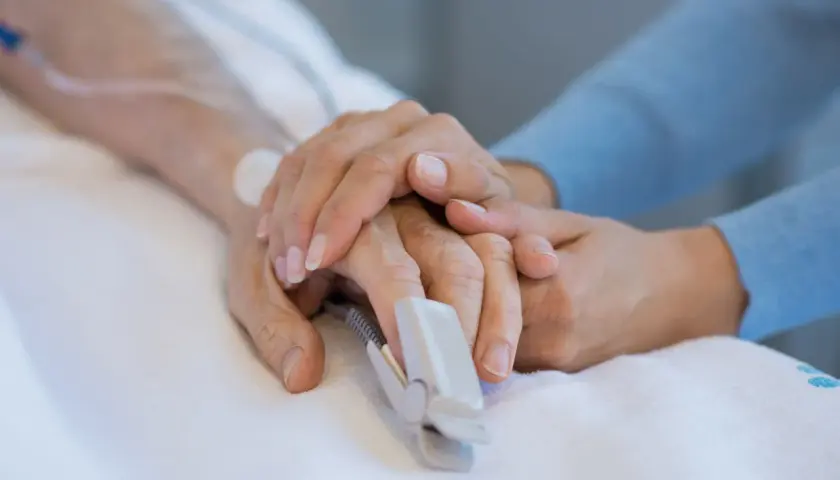 Dłoń pacjenta z pulsoksymetrem na palcu, otoczona troskliwymi dłońmi bliskiej osoby. To obraz nadziei i wsparcia na oddziale OIOM.