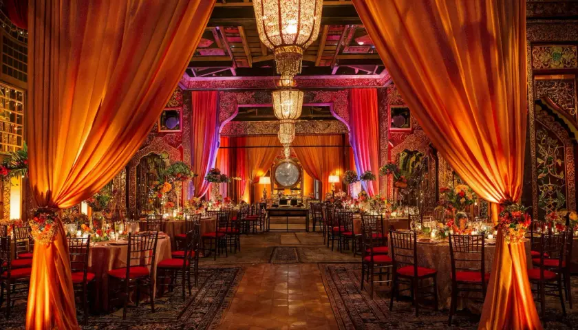 Décoration somptueuse pour un mariage bollywood, avec des drapés orangés, des lustres imposants et des tables dressées dans une ambiance chaleureuse et festive.