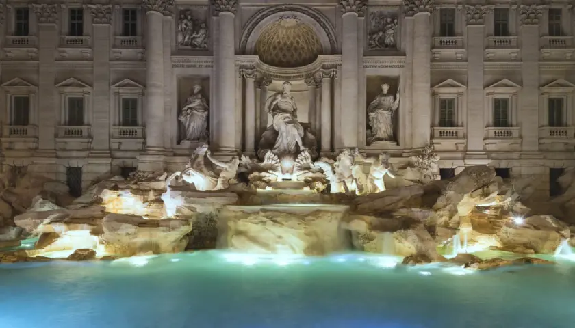 Fontana di Trevi stile barocco, con statue di divinità marine e cavalli marini che emergono da rocce e cascate, illuminata di notte.
