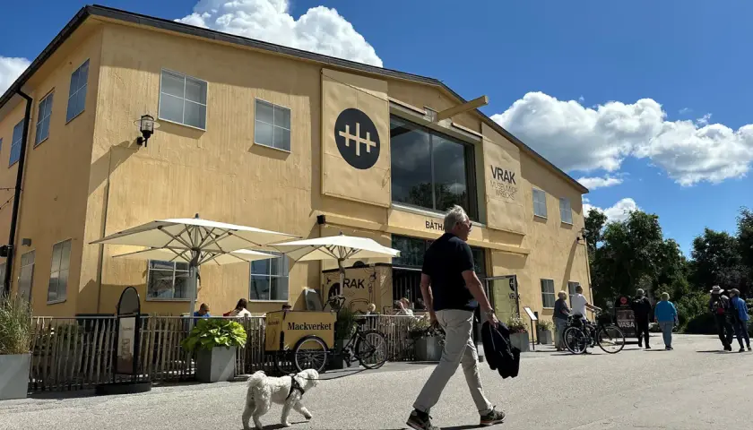 En gul byggnad med texten "VRAK MUSEUM OF WRECKS" under en stor svart symbol. Folk promenerar förbi, en hund går vid sidan av en man.