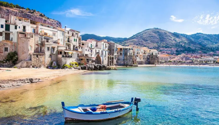 A charming fishing boat bobs in the clear turquoise water off the coast of Cefalù, a picturesque town with colorful buildings cascading down a hillside, making it one of the most beautiful places to go in Sicily.