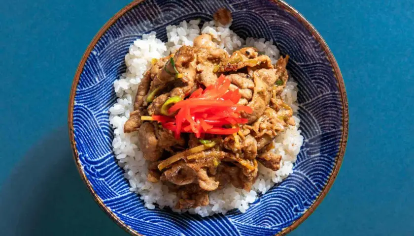 A bowl of fluffy white rice topped with savory pork shogayaki, garnished with bright red pickled ginger.